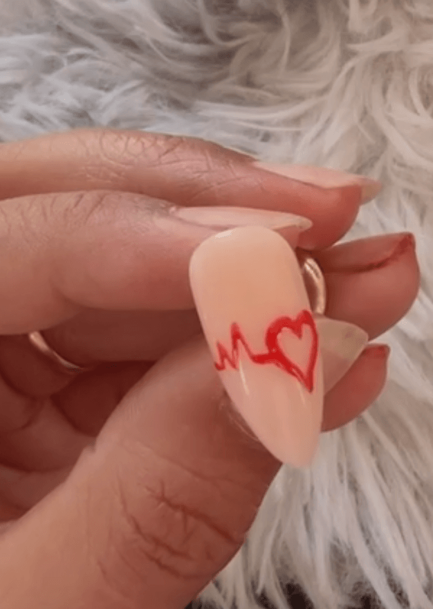 A close-up of the completed nail art design: a flawless, high-shine nude gel manicure featuring a clear red EKG heartbeat line flowing into a perfect red heart on the ring finger.