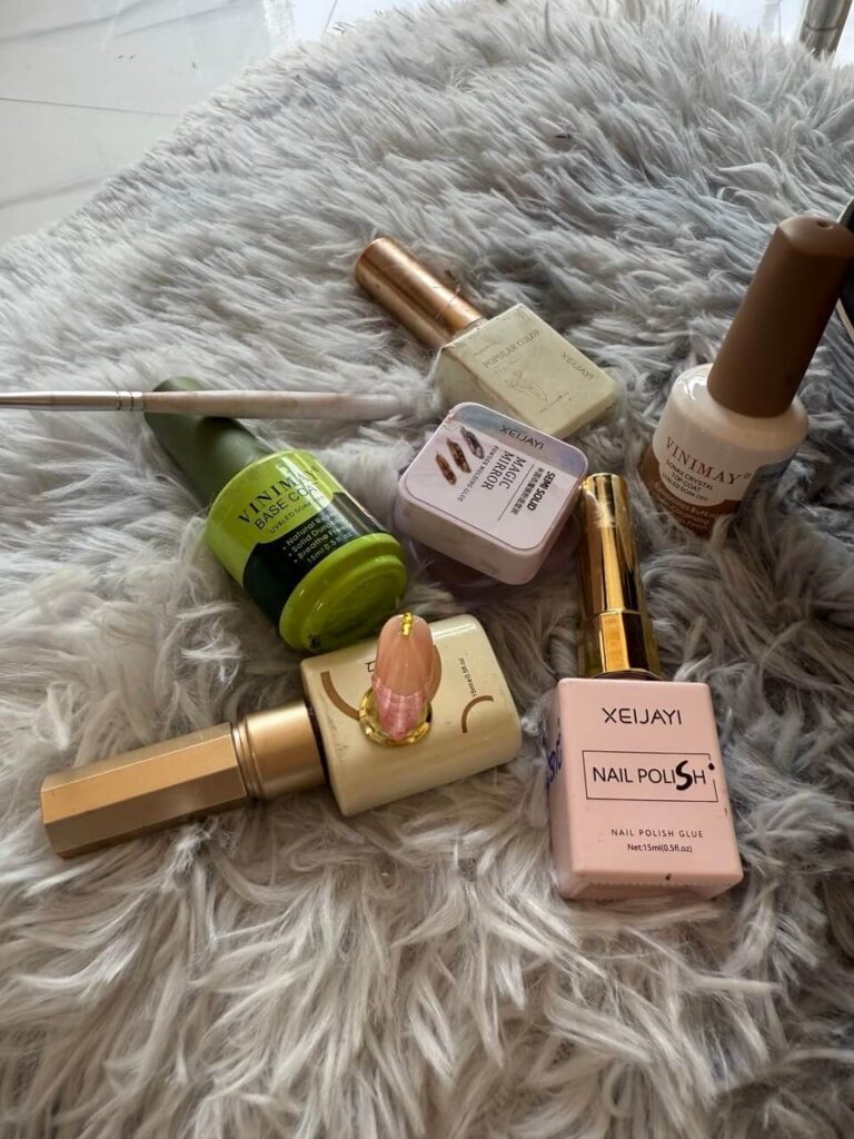 An overhead flat-lay photograph of various gel nail polishes, brushes, and a metal dotting tool arranged on a fluffy white surface.