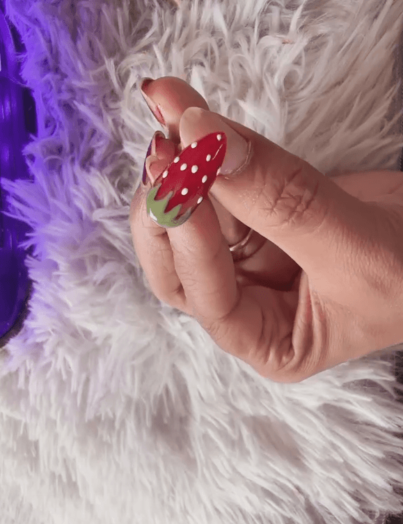 A close-up photograph of a finished strawberry nail art design, held up under the soft purple light of a UV/LED nail lamp.