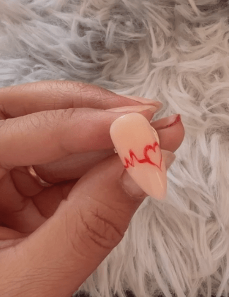 Close-up showing the hand holding the nail form after painting the wave, now transitioning the liner brush to outline the curve of a red heart shape.