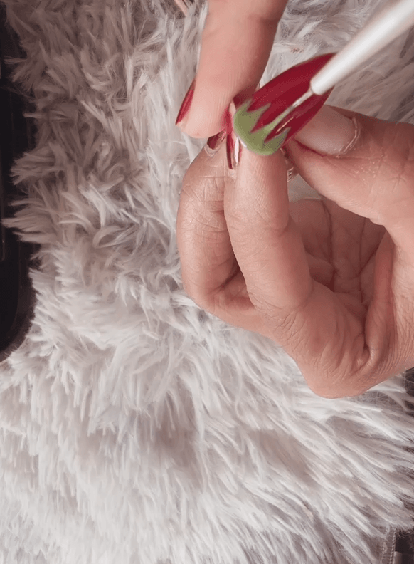 A close-up shot showing a fine-tip liner brush actively painting green gel leaves onto the base of a red nail.