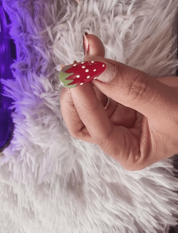 A close-up photograph showing a person holding up an almond-shaped nail with completed red strawberry nail art, including green leaves and white dots.